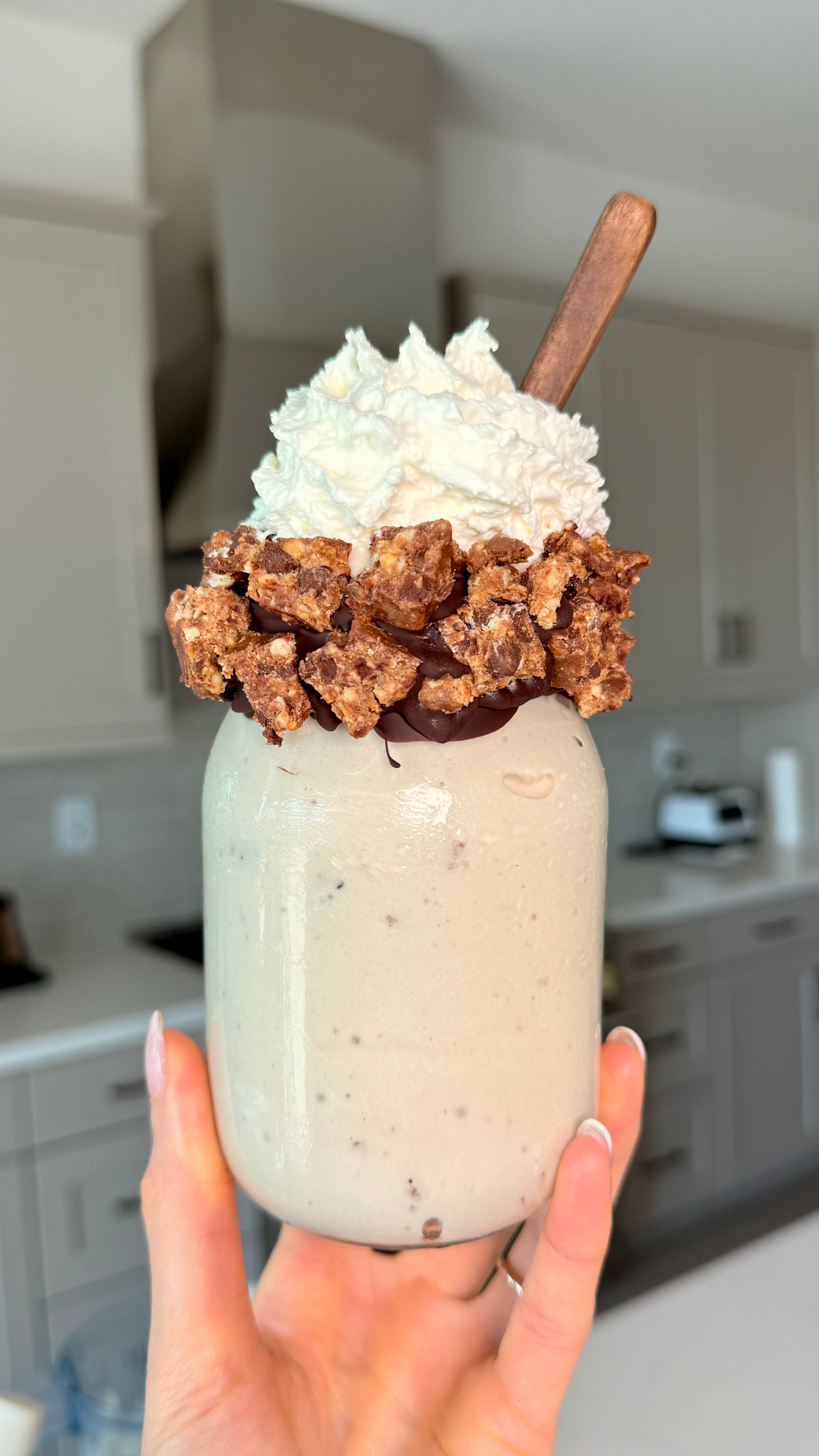 Hand holding a jar of creamy dessert with whipped cream and a cinnamon stick in a kitchen setting